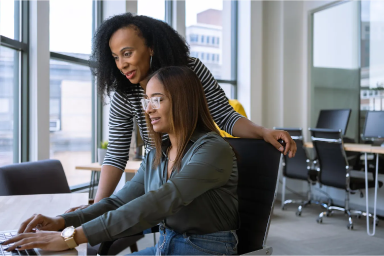 A supportive mentorship scene where an experienced woman (mentor) is standing behind a younger woman (mentee) who is wearing glasses and working intently on a laptop in a modern office. The mentor is leaning in, smiling, and placing a hand on the mentee's chair/shoulder, guiding her work.